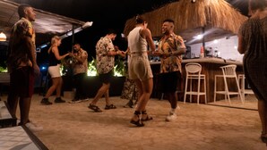 Un grupo de personas bailando en un espacio al aire libre con una estructura de techo de paja y sillas blancas.