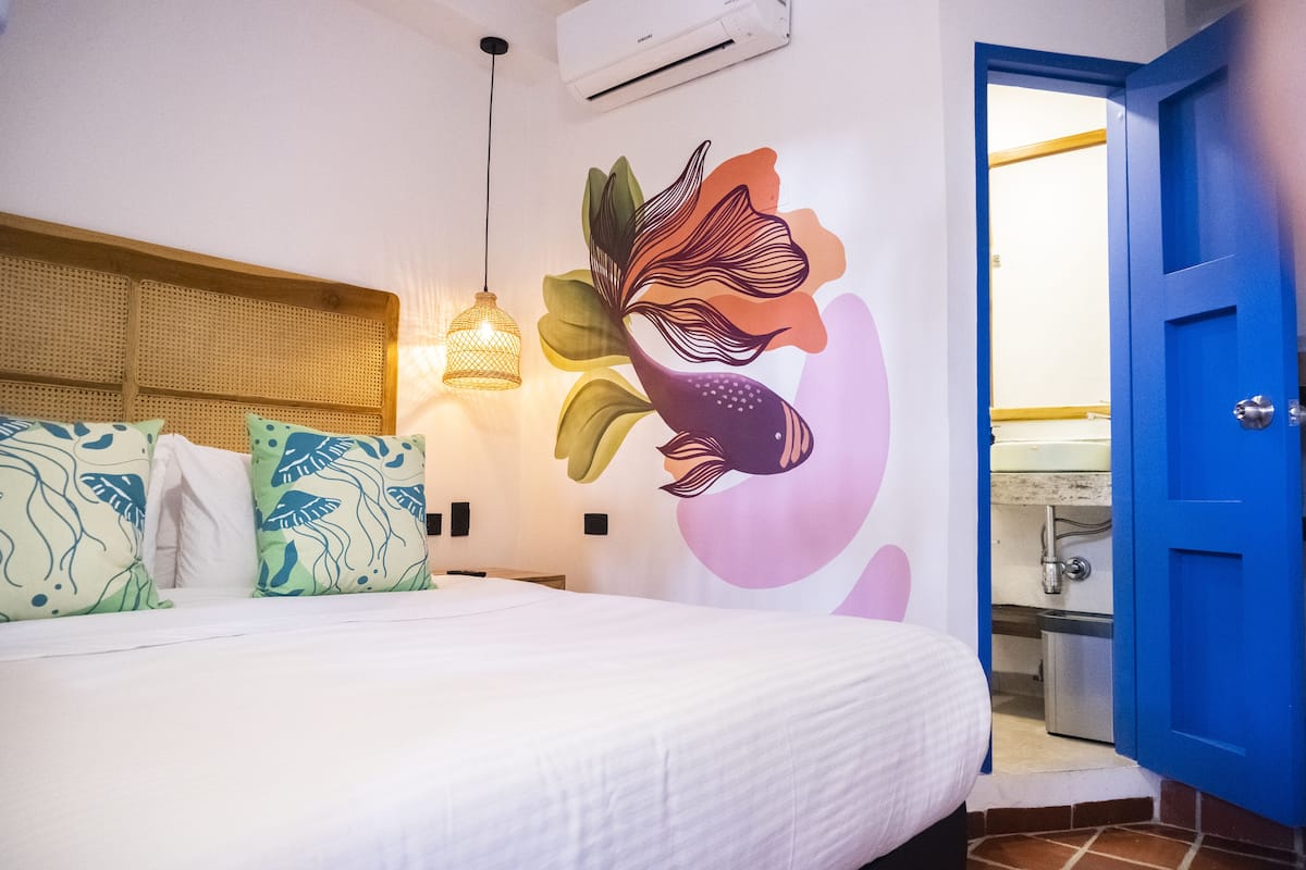 A bedroom with a bed, decorative pillows, a wall mural, a hanging light fixture, and a blue door leading to a bathroom.