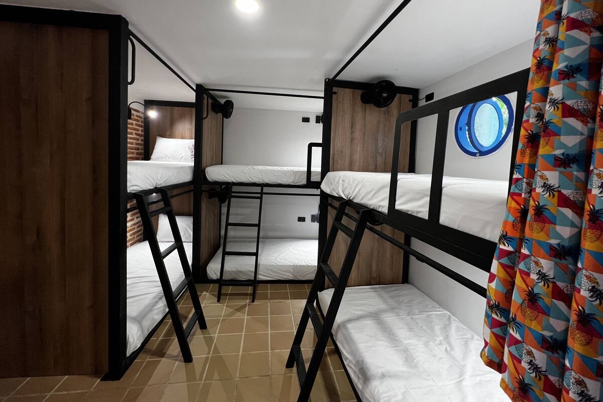 A bunk bed room with a wooden door, a window with a blue frame, and a colorful curtain.