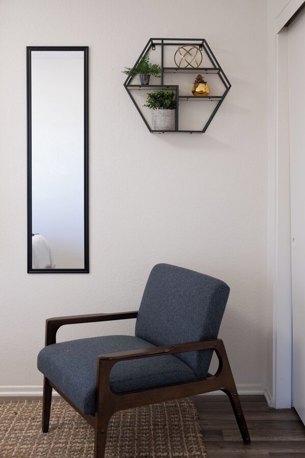 Una silla con estructura de madera oscura y asiento de tela azul, ubicada frente a un espejo montado en la pared y un estante geométrico con plantas y objetos decorativos.