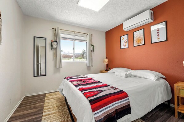 A hotel room with a bed, a window with curtains, a wall-mounted air conditioner, and framed artwork.
