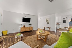 A modern living room with a flat-screen TV, a dining area with a round table, and a kitchen in the background.