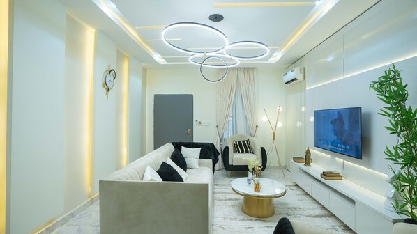 Un salon moderne avec un canapé, un fauteuil et une table basse. Un téléviseur est fixé au mur et une plante se trouve sur la droite.