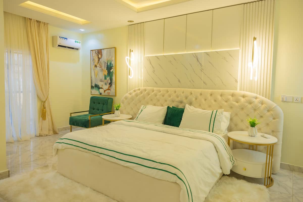 A modern bedroom with a large bed, a green armchair, a small round table, and a wall-mounted air conditioner.