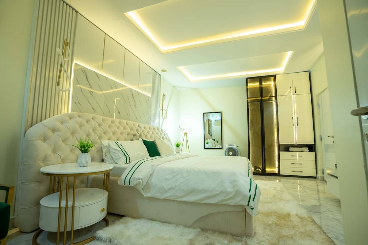 A modern bedroom with a tufted headboard, a round bedside table, a fluffy rug, and built-in wardrobe with LED lighting.