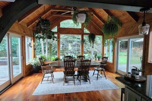 A dining area with a wooden table and chairs, surrounded by large windows and hanging plants. - Exquisite cottage with valley views and Hot Tub (New Holland)
