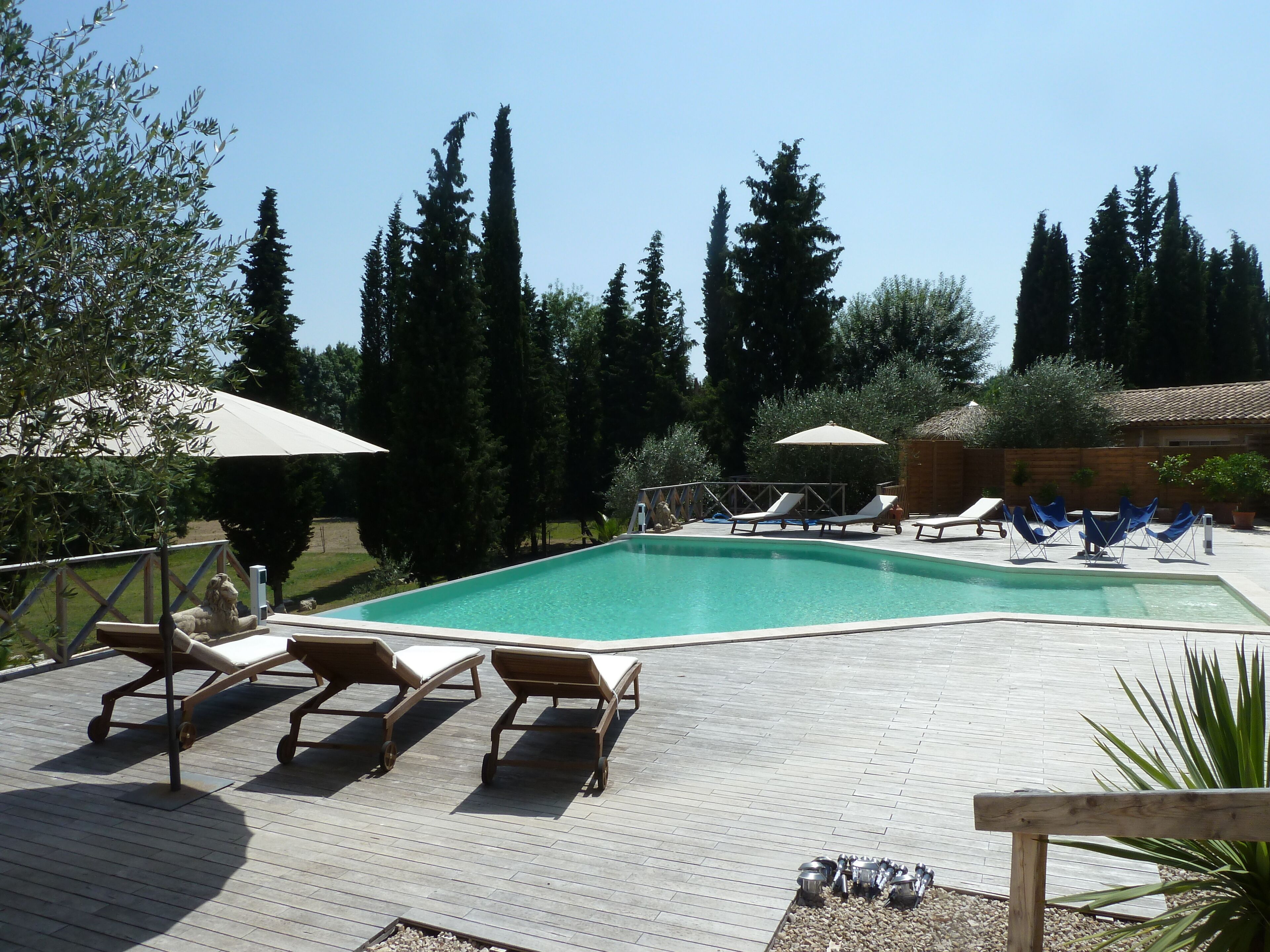 Une piscine entourée de chaises longues et de parasols, bordée de grands arbres et d’une terrasse en bois.
