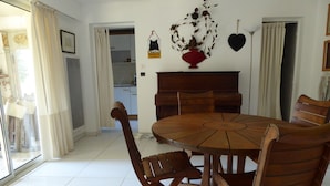 A dining area with a round wooden table and chairs, a kitchen in the background, and a decorative wall piece. - Charming cocoon for 4p in nature, space, infinity pool (Châteauneuf-Grasse)