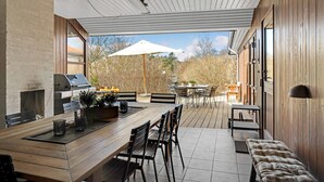 Eine überdachte Terrasse mit Holztisch und -stühlen, einem Grill und Blick auf den Garten.