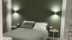 Un dormitorio con pared verde, una cama con colcha verde, dos mesitas de noche con libros y una lámpara, y una lámpara de techo blanca.
