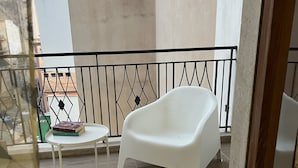 Un balcón con una silla de plástico blanca, una mesita redonda con libros y una barandilla de metal.