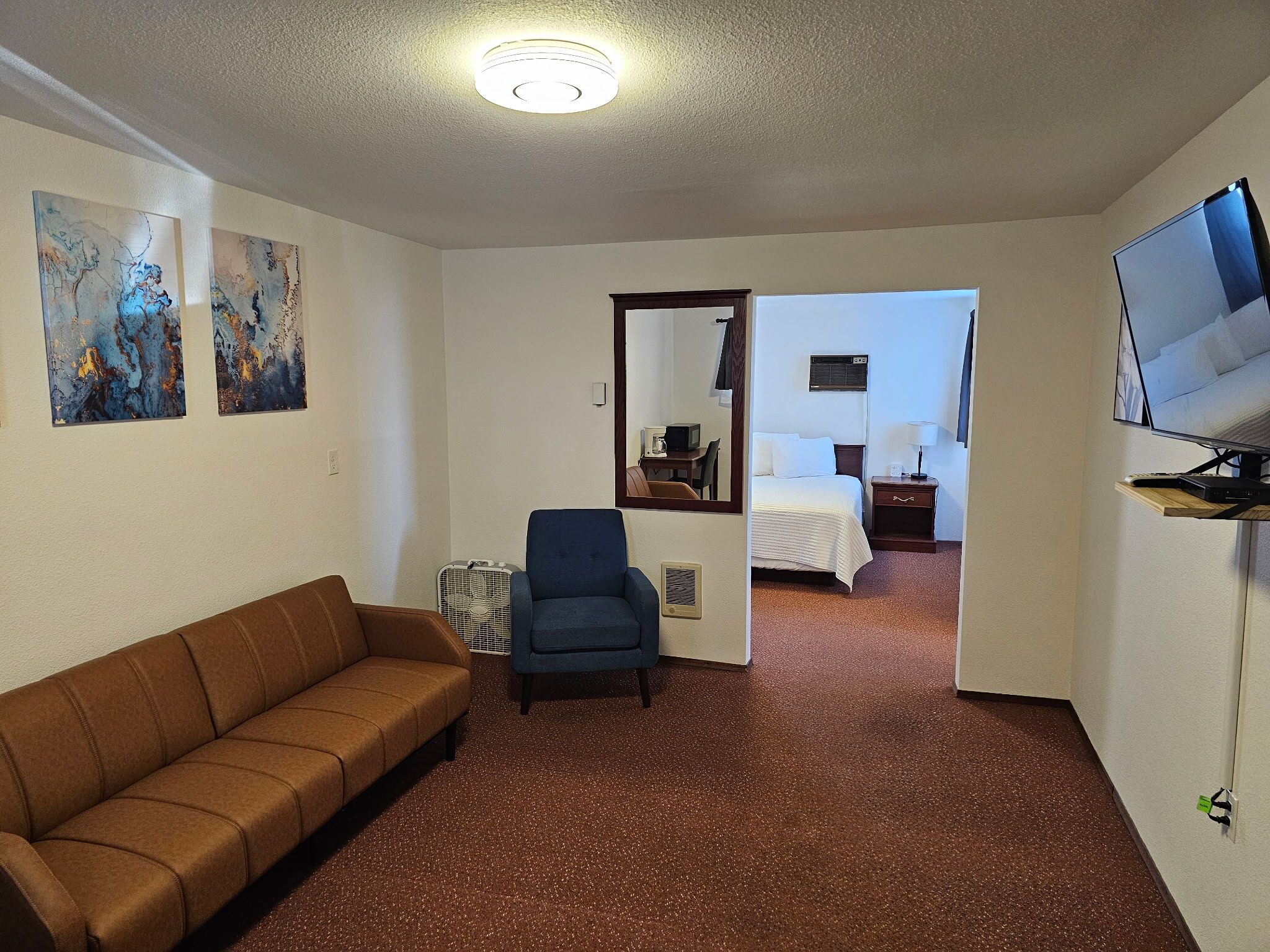 Een hotelkamer met een bruine bank, een blauwe fauteuil, een bed, een televisie en abstracte kunst aan de muren.