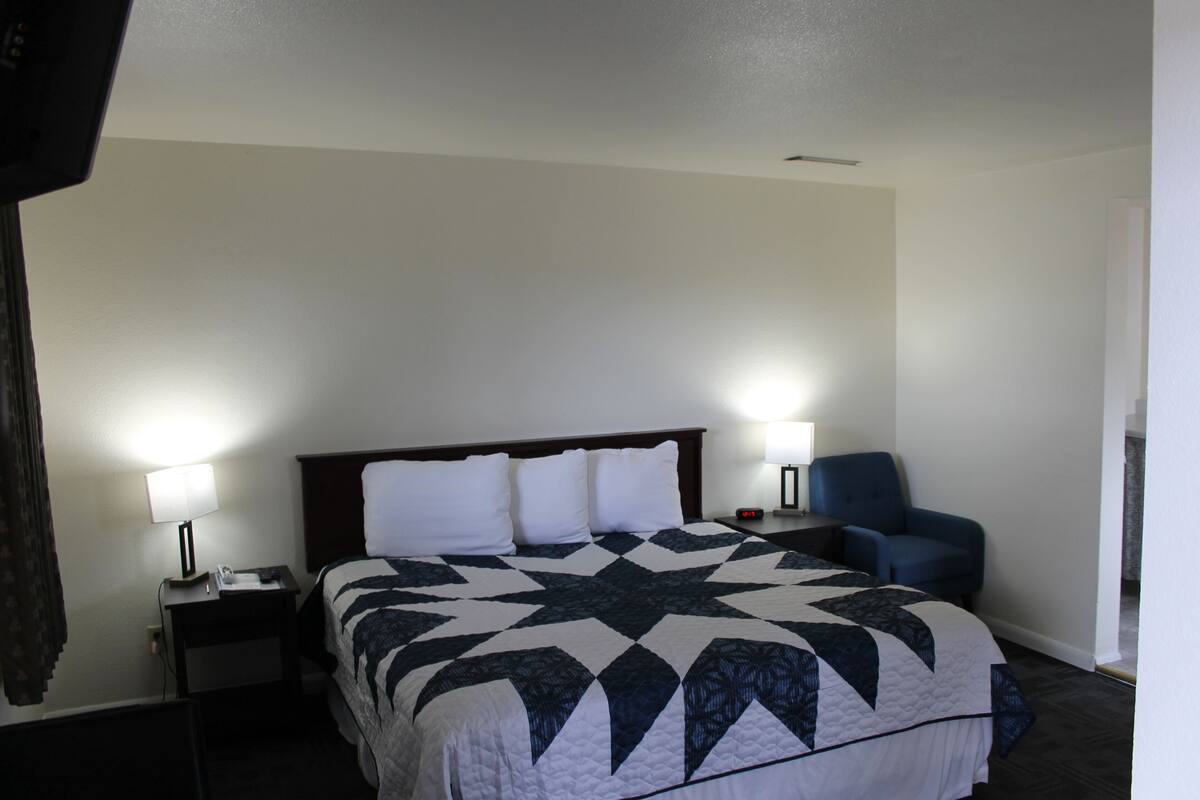 A hotel room with a large bed, two bedside lamps, a chair, and a nightstand with a phone and a book.