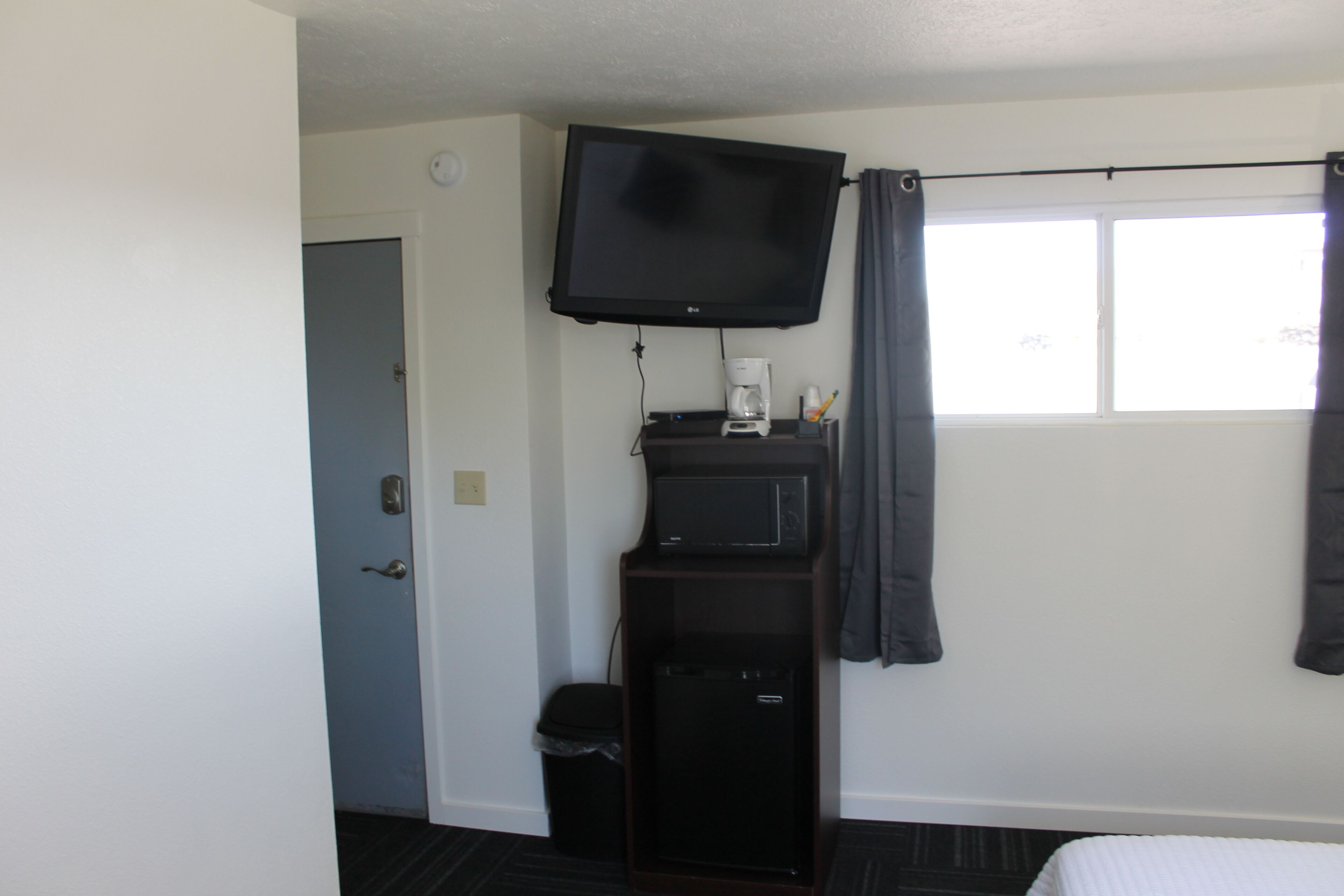 A hotel room with a flat-screen TV mounted on the wall, a small refrigerator, a trash bin, and a window with curtains.