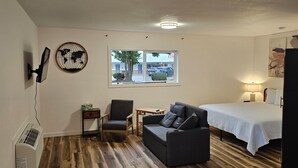 Una habitación con una cama, un sofá gris, una mesita, una silla, una obra de arte con un mapa mundial, una ventana que ofrece vistas a edificios y coches, y una lámpara en el techo.