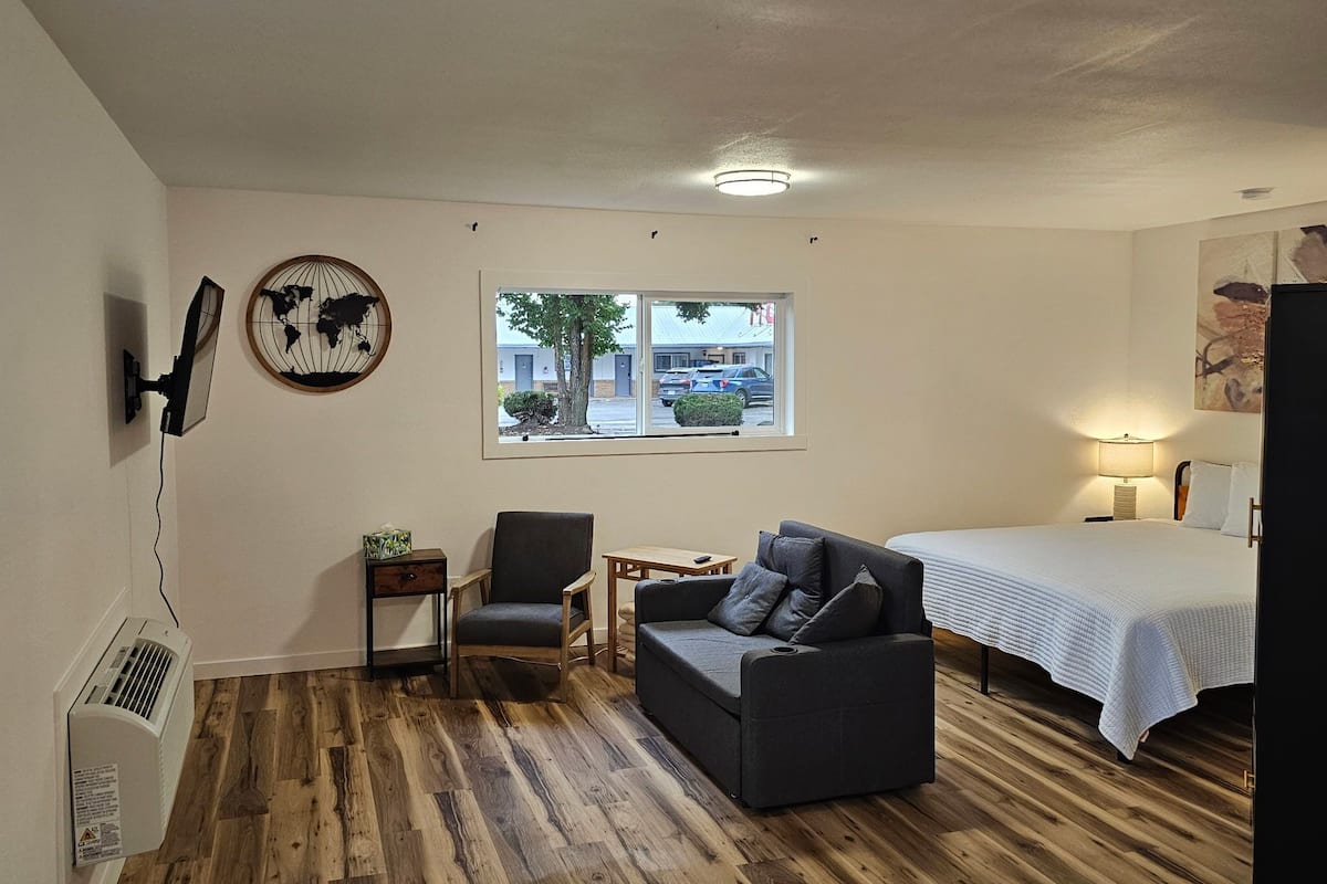 A room with a bed, a grey sofa, a small table, a chair, a world map artwork, a window with a view of buildings and cars, and a ceiling light.