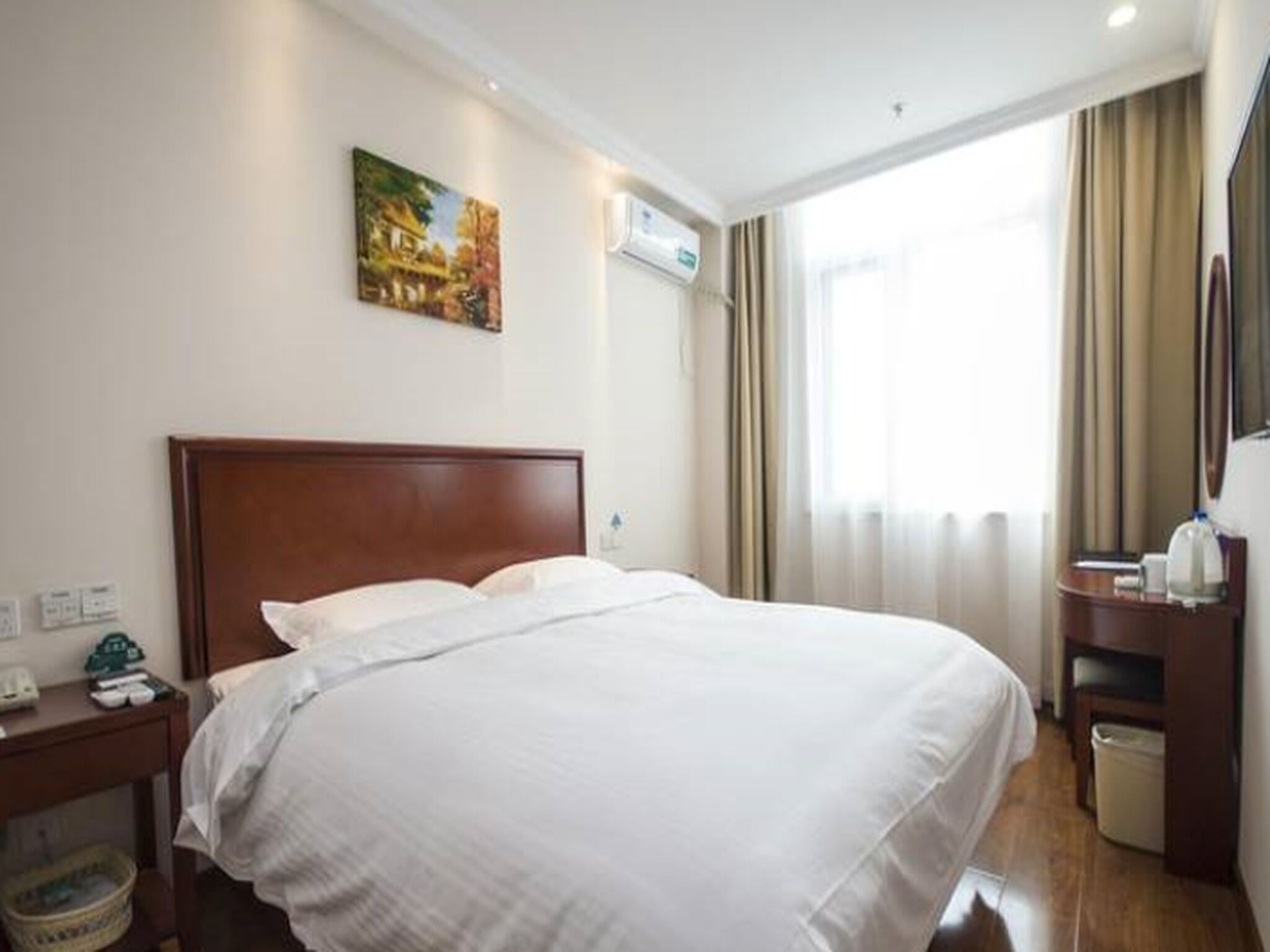 Habitación de hotel con una cama grande, una mesita de noche con lámpara, un cesto de basura pequeño y un cuadro en la pared.