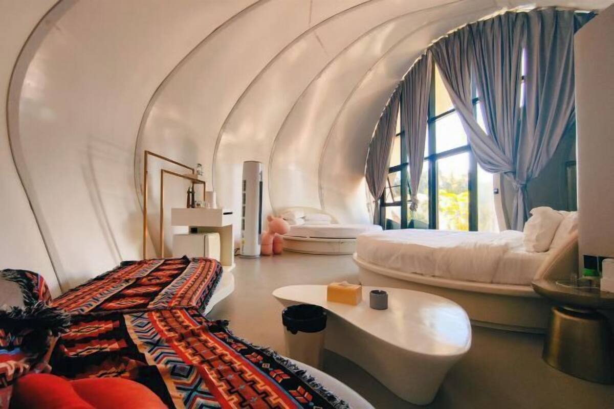 A modern, curved room with a bed, a sofa, a coffee table, and a large window with curtains.