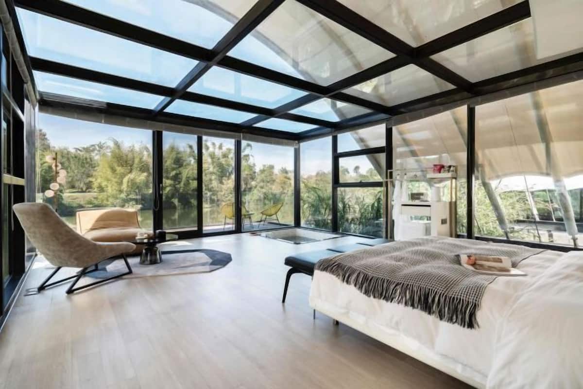 A modern bedroom with a large bed, a chair, and a desk. It features large windows and a view of the outdoors.