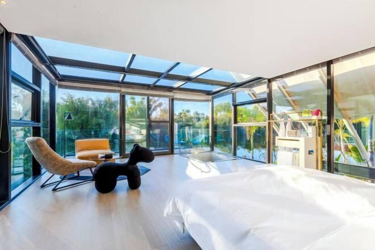 A modern bedroom with a large bed, a sofa, and a chair, with a glass wall offering a view of greenery.
