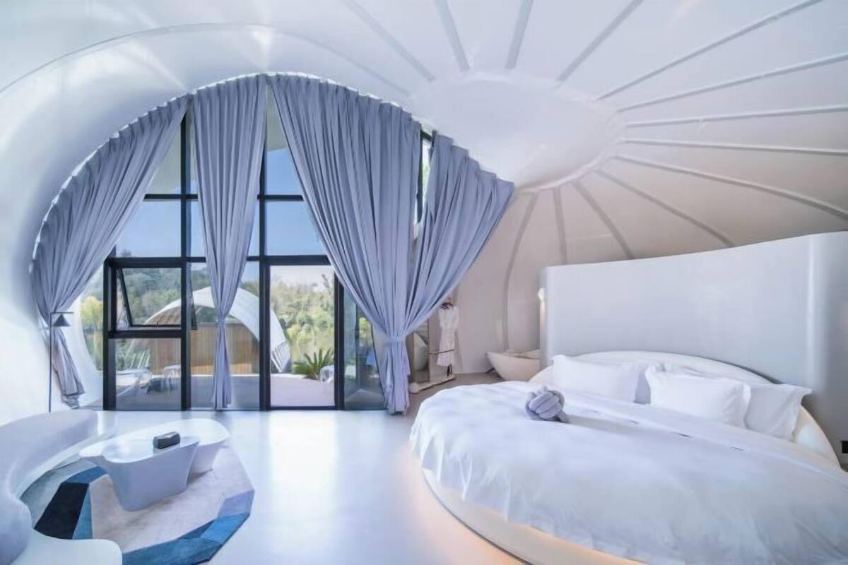 A modern bedroom with a large bed, a curved seating area, and a view through a large window.