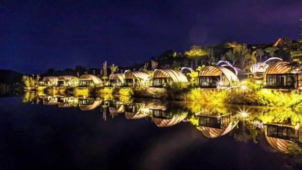 A series of circular, boat-like structures with reflective surfaces, situated on a calm body of water, reflecting the structures and surrounding greenery. - Xishuangbanna Visible Zeqi Tent Hotel (Menghai)
