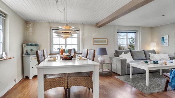 A modern living room with a dining area, featuring a white ceiling, wooden beams, a large window, and a grey sofa. - Lovely home in Pandrup with WiFi (Pandrup)