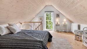 A cozy attic bedroom with a sloped ceiling, a large bed with a black quilt, a wicker chair, and a view of greenery through a window. - Lovely home in Pandrup with WiFi (Pandrup)