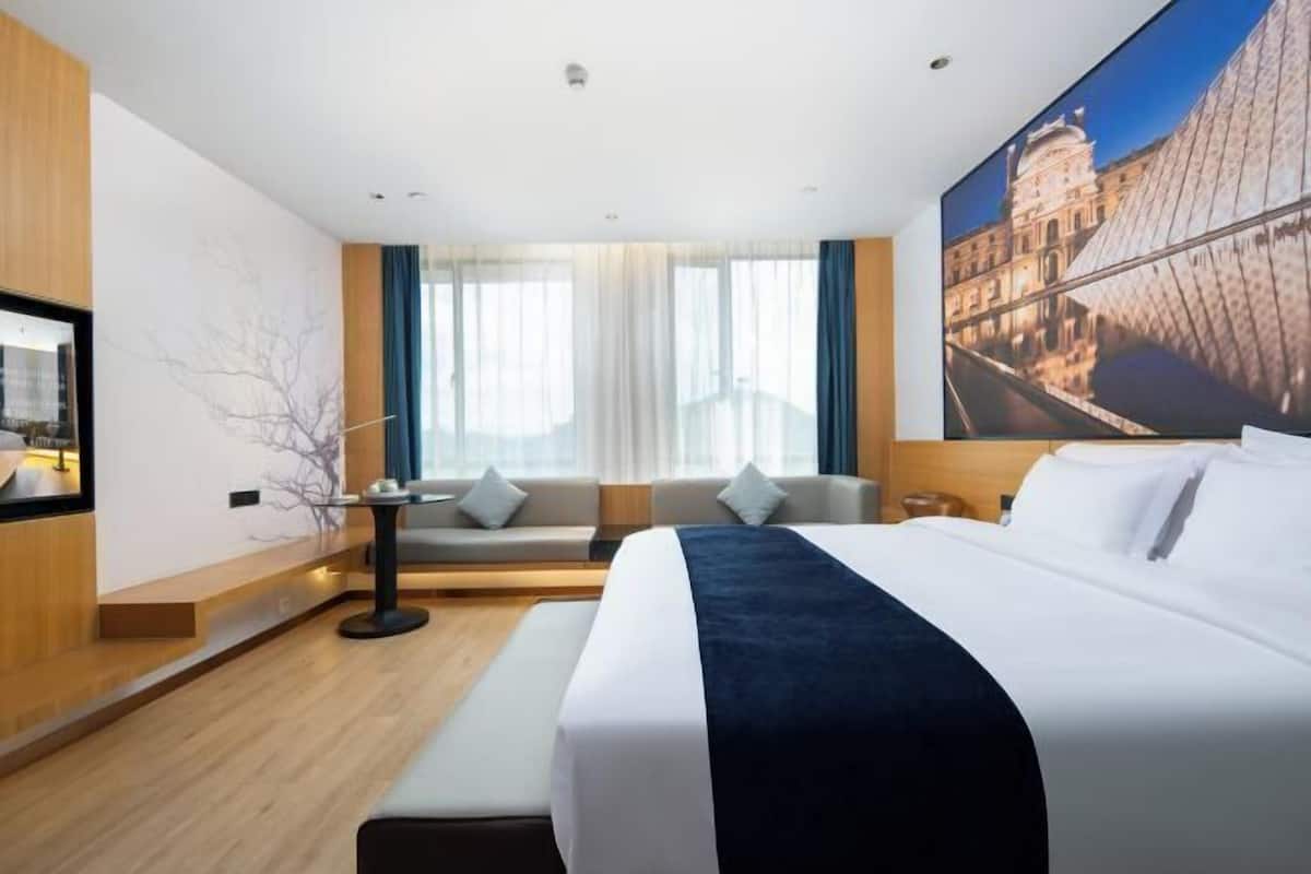 Ein modernes Hotelzimmer mit einem großen Bett, einer Sitzecke mit einer Couch und Sesseln, einem kleinen Tisch und einem großen Fenster mit Vorhängen.