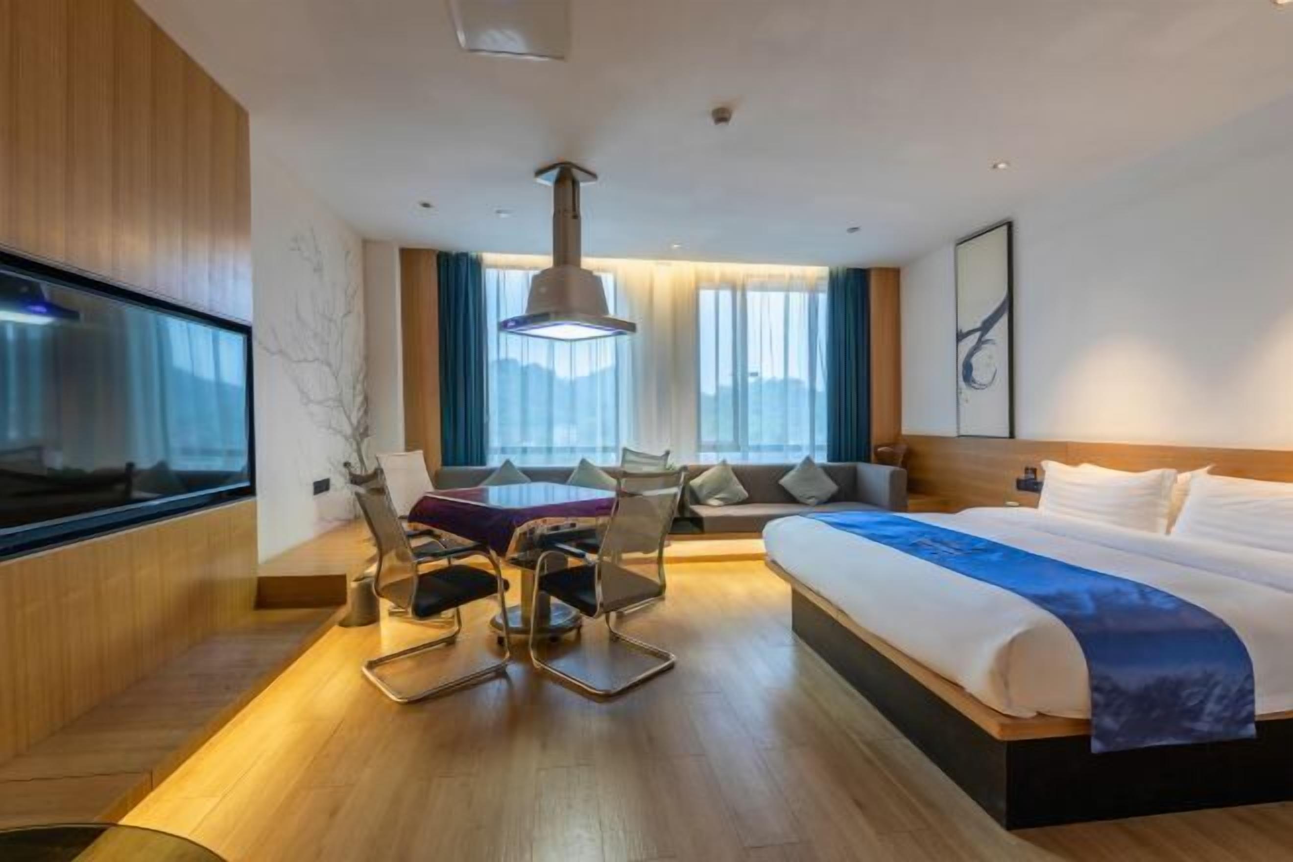 A modern hotel room with a large bed, a dining table with chairs, a TV, and a large window with curtains.