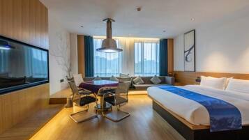 Habitación de hotel moderna con una cama grande, una mesa de comedor con sillas, un televisor y un ventanal con cortinas.