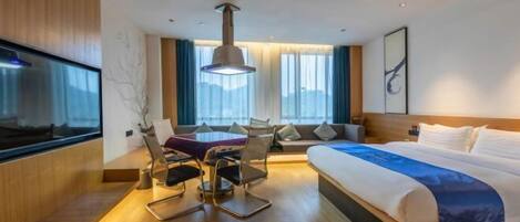 A modern hotel room with a large bed, a dining table with chairs, a TV, and a large window with curtains.