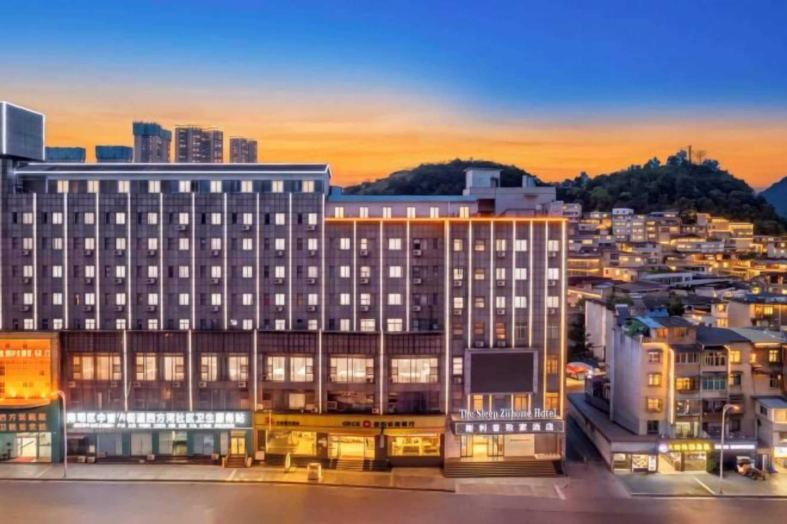 A multi-story hotel with a modern facade, located in an urban setting with a view of the cityscape and a hill in the background.
