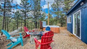 A backyard with a stone fire pit, two Adirondack chairs, a hot tub, and a blue building with a glass door. - Cabin in the Woods with Hot Tub, Incredible Views, Fireplace, Foosball, Fire Pit (Green Mountain Falls)