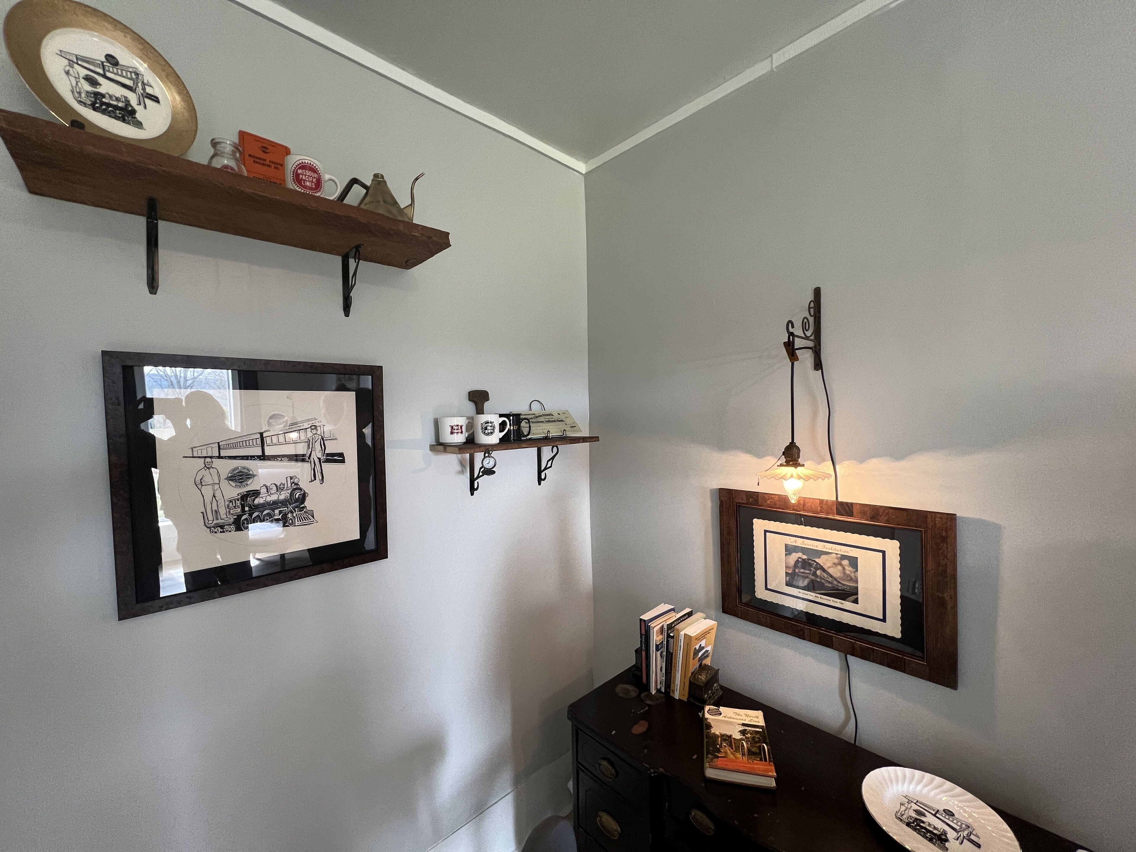 Una habitación con un cuadro de un tren, una estantería con tazas y una lámpara fijada en la pared.