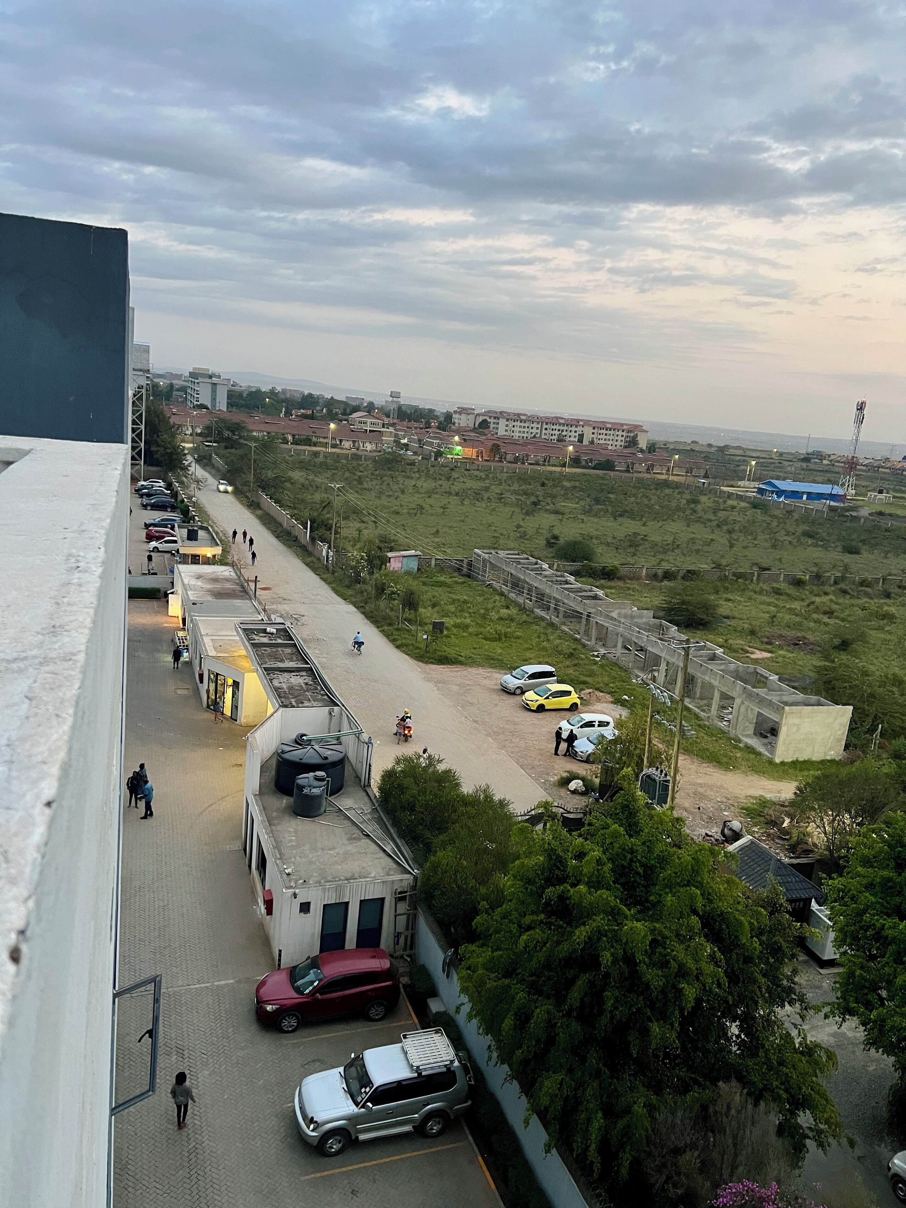 Vista de un estacionamiento con varios coches y un autobús, un edificio de techo plano, y una vista distante de una zona residencial con casas y un torre de agua.