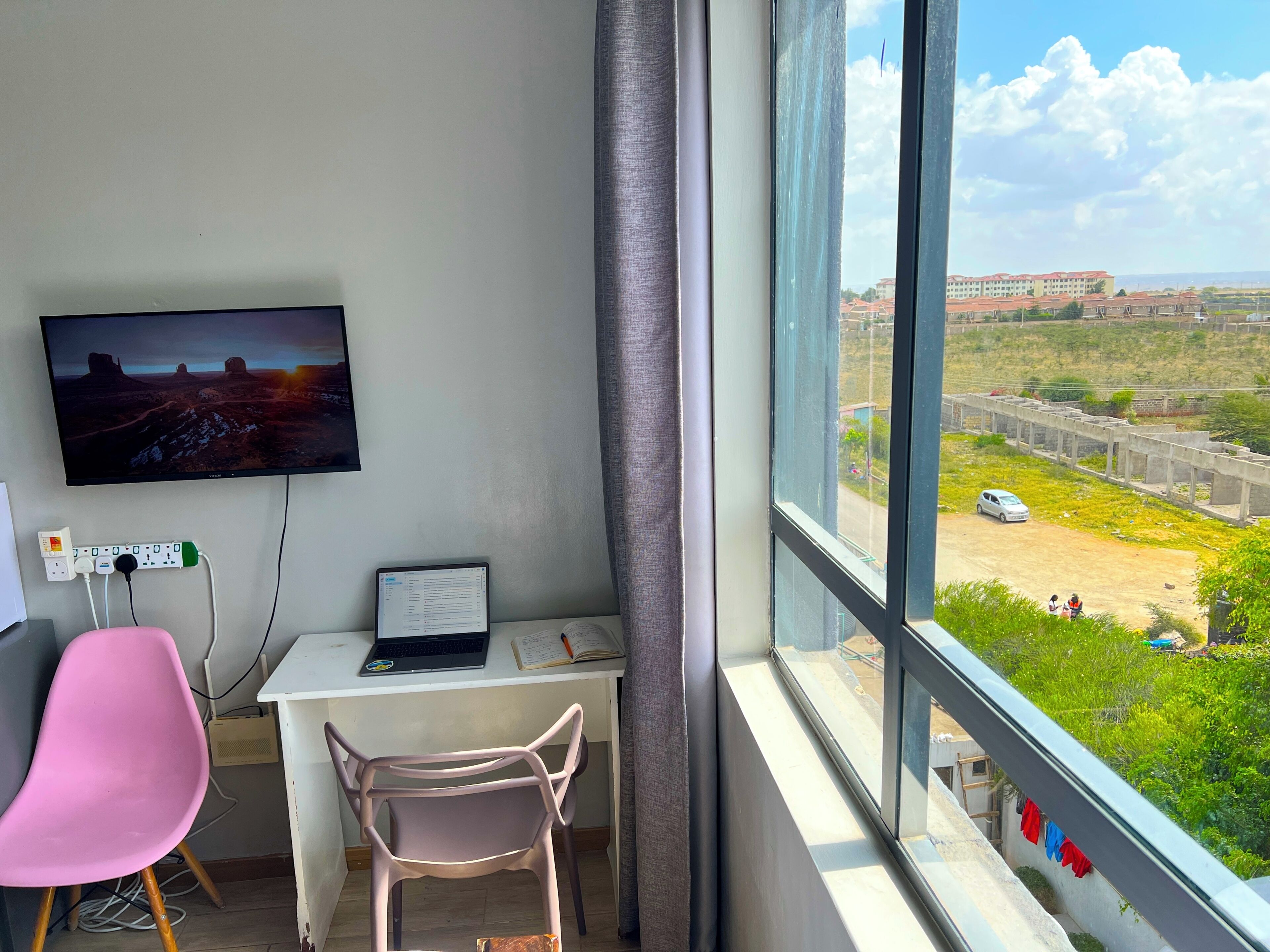 A room with a desk, a pink chair, a laptop, a TV on the wall, and a large window with a view of a building and greenery.
