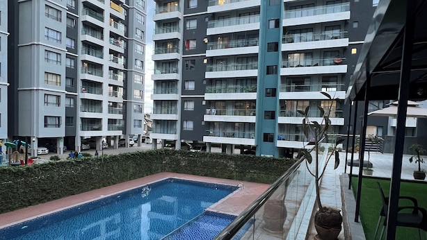 A modern apartment building with a rectangular swimming pool and a covered outdoor area with chairs and potted plants. - Indesign Studio  (Syokimau)