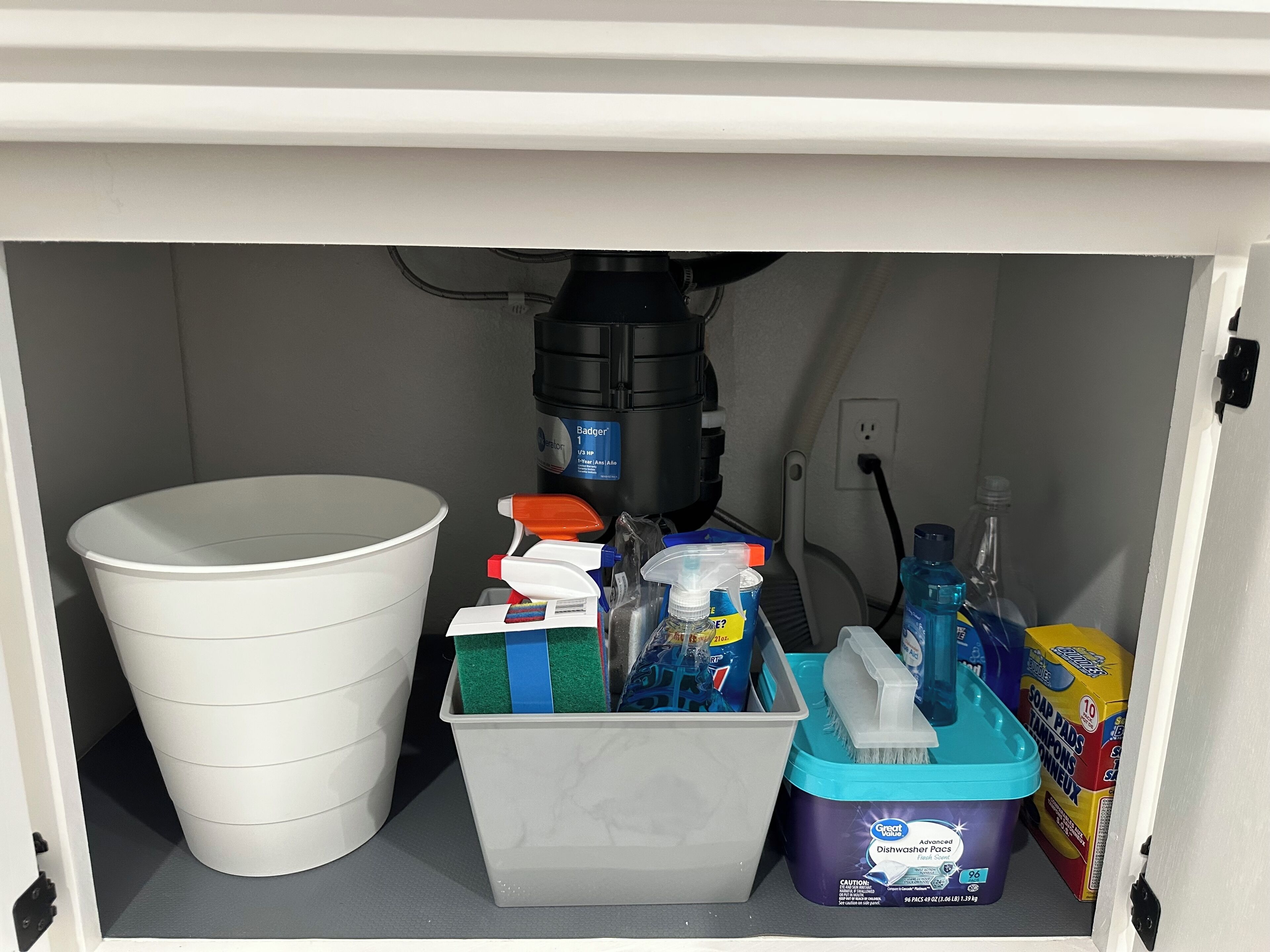 A bathroom with a white toilet, a white bucket, a plastic container with cleaning supplies, a blue dishwashing soap container, and a yellow box of soap pads.