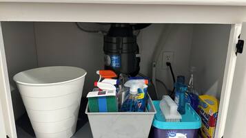 A bathroom with a white toilet, a white bucket, a plastic container with cleaning supplies, a blue dishwashing soap container, and a yellow box of soap pads.