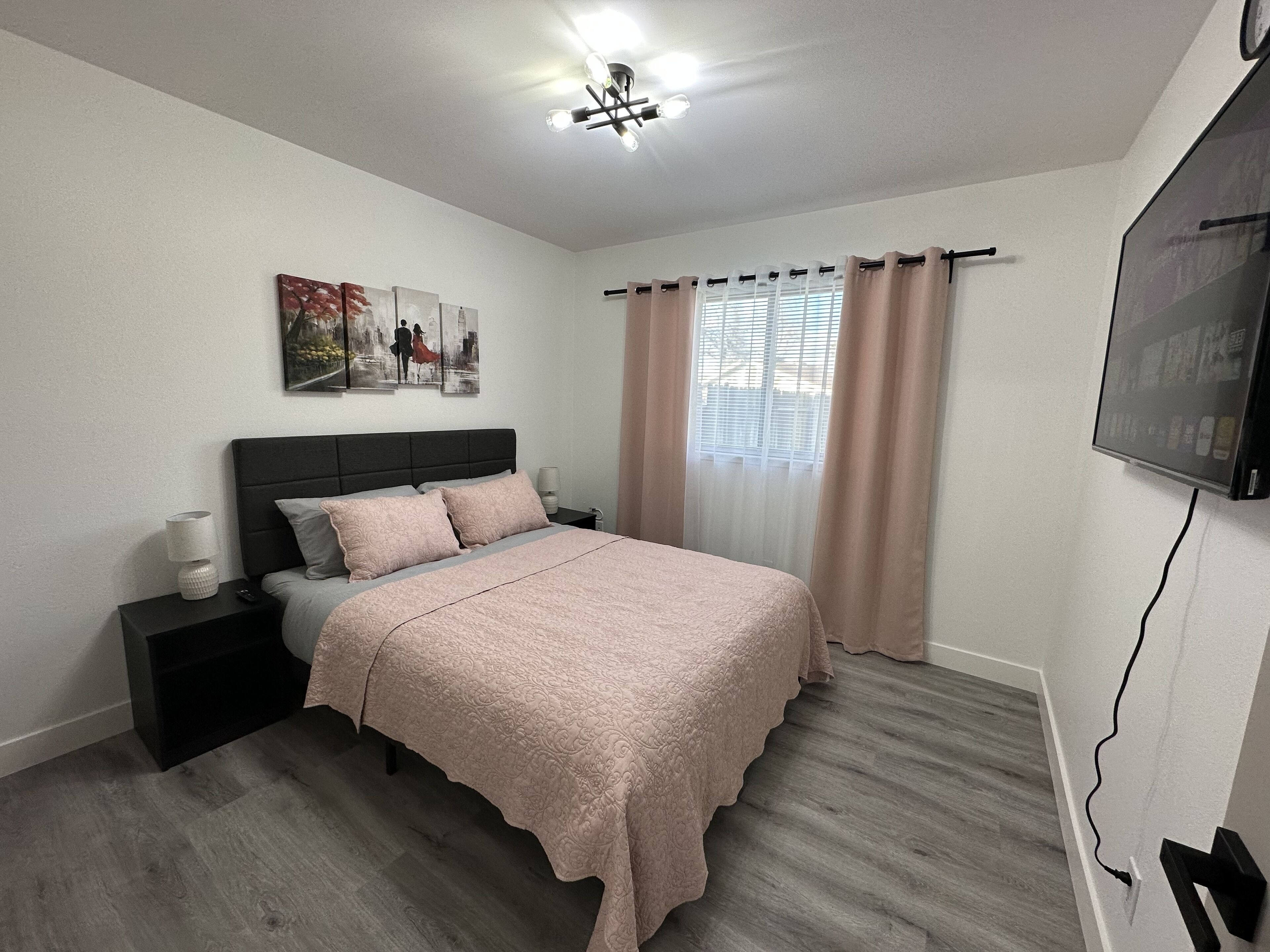 Une chambre à coucher comprenant un lit, des tables de chevet, une lampe, un tableau, une fenêtre avec des rideaux et un téléviseur fixé au mur.