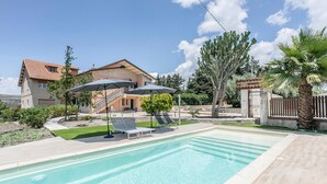 Una piscina con sillas de descanso y sombrillas, rodeada de un jardín ajardinado y una casa con tejado de tejas.
