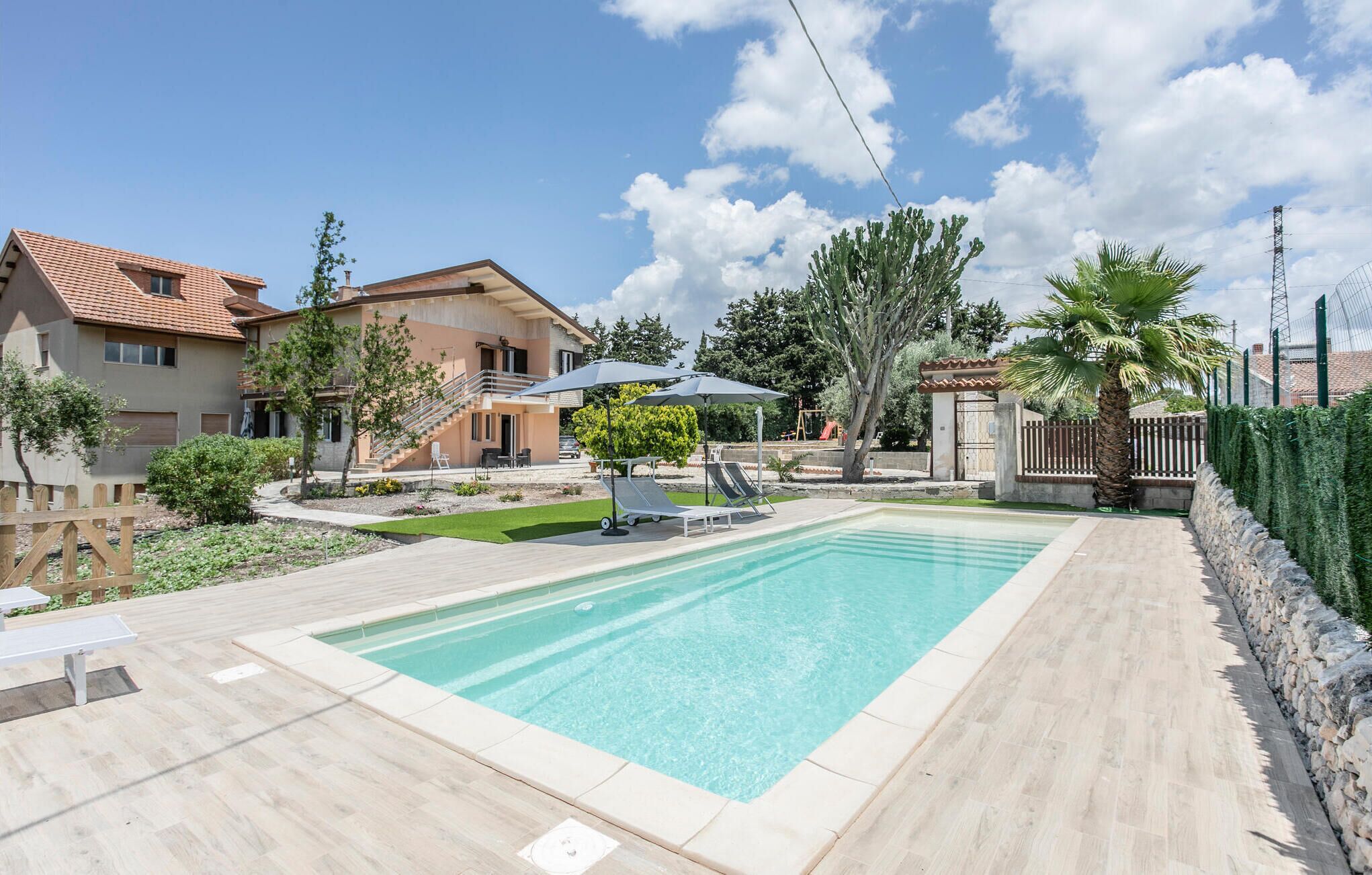 A swimming pool with a wooden deck, a covered patio area, and a house with a red-tiled roof.