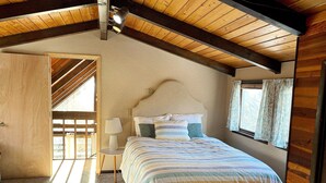 Un dormitorio con cama, mesita de noche, lámpara, ventana con cortinas y techo de madera.