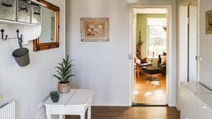 Un pasillo con una mesa blanca, una planta en maceta, un cuadro enmarcado y una puerta que da a una habitación con una silla y una lámpara.