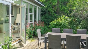 Un patio con mobiliario de mimbre, un ventanal de vidrio que da a una habitación y abundante follaje.