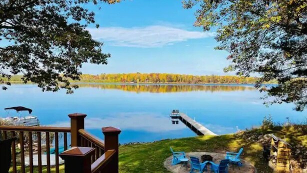 Ein Seeufer mit Terrasse und Blick auf einen ruhigen See, eine Anlegestelle und Herbstlaub.