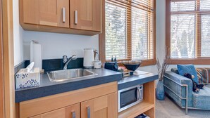 A modern kitchen with wooden cabinets, a stainless steel sink, and a built-in microwave. There is a window with blinds, a sofa with a teddy bear, and a vase with branches. - A Peaceful Mountain Retreat in one of Banff's Most Elegant Homes (Banff)