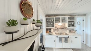 Une cuisine moderne avec des meubles blancs, un four encastré et un évier en acier inoxydable. Des plantes en pot sont disposées sur une étagère au-dessus du plan de travail, et un panneau décoratif orne le mur.