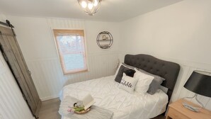 Un lit bien fait, avec une couette et des oreillers blancs, une table de chevet avec une lampe, une étagère murale ornée d’objets décoratifs et une fenêtre avec des stores.